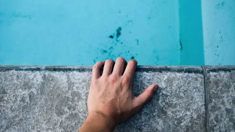 Main posée sur la margelle d'une piscine dont le fond présente des taches sombres