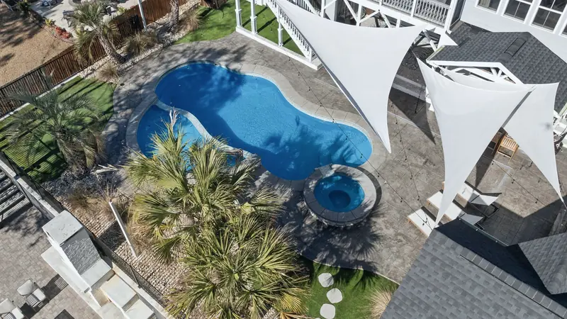 Vue aérienne d'une piscine enterrée aux formes arrondies et d'un spa rond, entourés de palmiers et de voiles d'ombrage