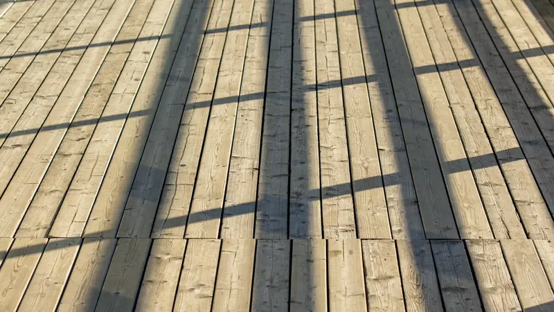Platelage en lames de bois claires aboutées sur plusieurs rangs, rayé par les ombres d'une rambarde au soleil rasant