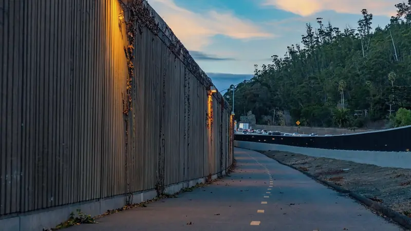 Haut mur antibruit en béton rainuré éclairé au crépuscule, longé par une piste cyclable, avec une voie rapide chargée de voitures en arrière-plan