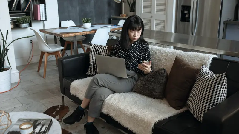 Femme assise sur un canapé, un ordinateur portable sur les genoux, consultant son smartphone
