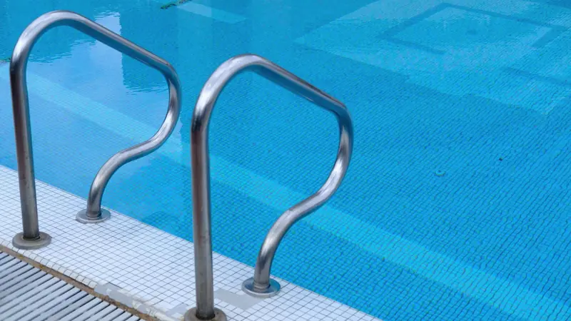 Main courante en inox double fixée sur une margelle carrelée, au bord d'un bassin à mosaïque bleue