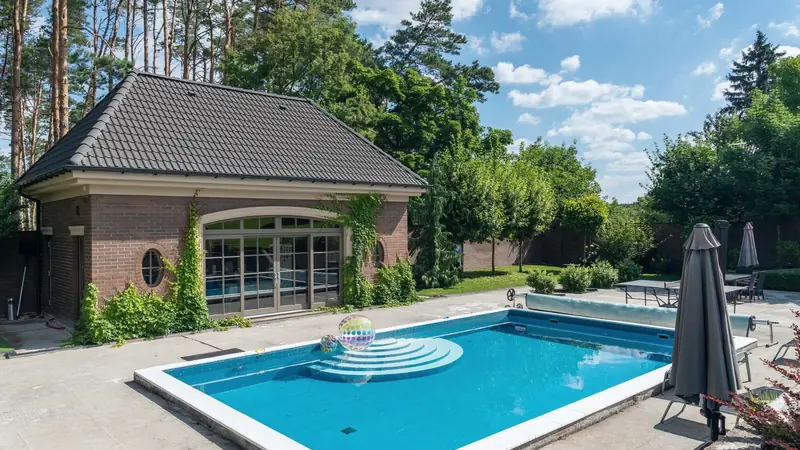 Piscine enterrée rectangulaire avec escalier roman arrondi, couverture enroulée en bout de bassin et pool house en brique au fond du jardin