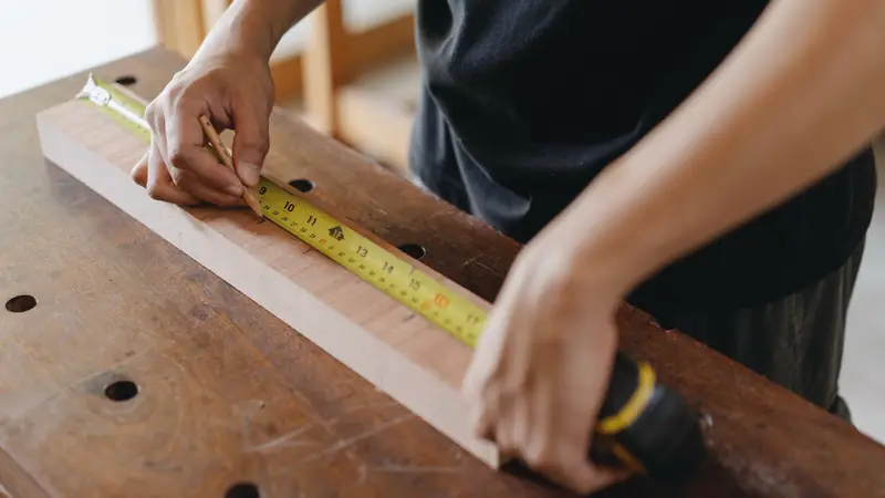 Mains traçant un repère au crayon sur une pièce de bois, le long d'un mètre ruban jaune posé sur un établi