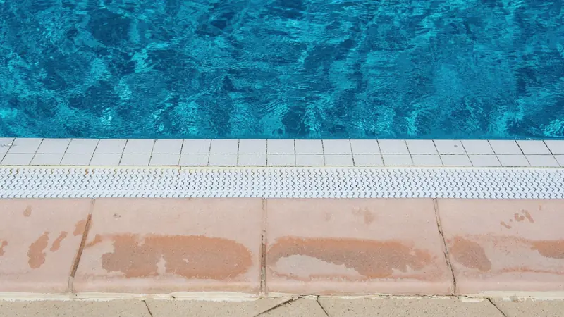 Bord de piscine carrelé avec margelles, grille de goulotte et eau turquoise agitée par le vent