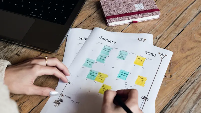 Mains annotant au feutre un calendrier mensuel imprimé posé près d'un ordinateur portable et d'un agenda