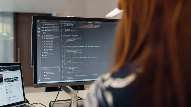 Développeuse vue de dos devant un écran ouvert sur un éditeur de code, un portable à côté