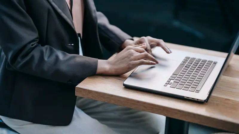 Mains d'une personne en veste sombre tapant sur le clavier d'un ordinateur portable posé sur une table en bois